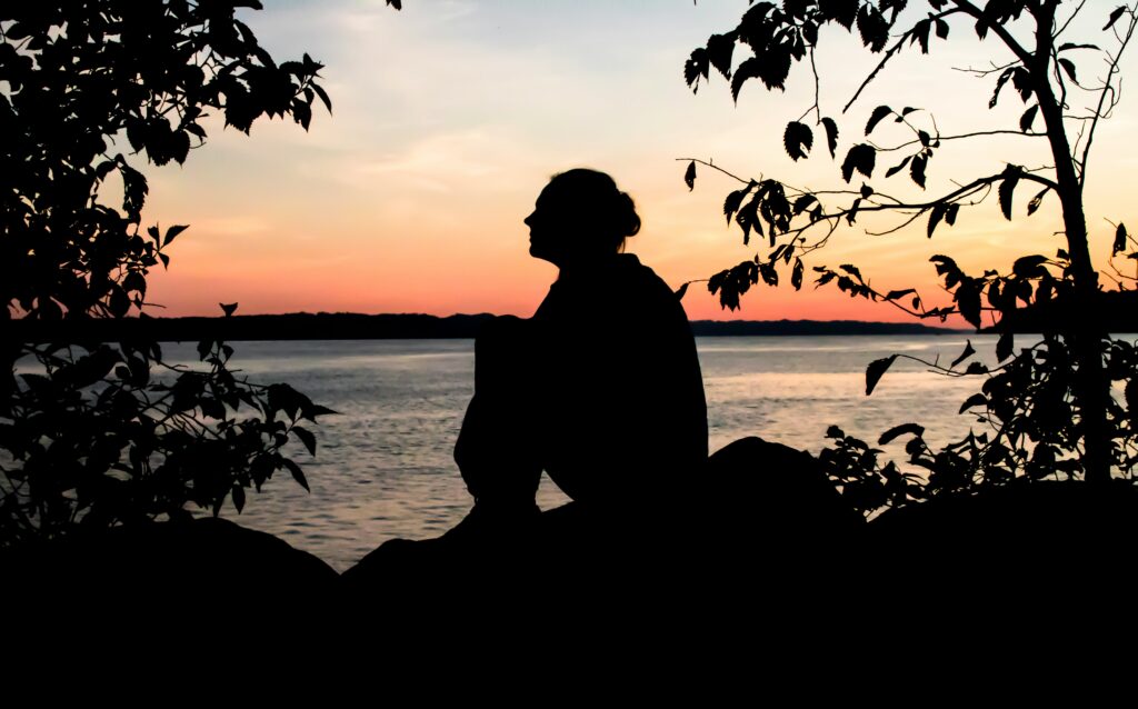 Person sitzt am Seeufer bei Sonnenuntergang und blickt auf das Wasser.